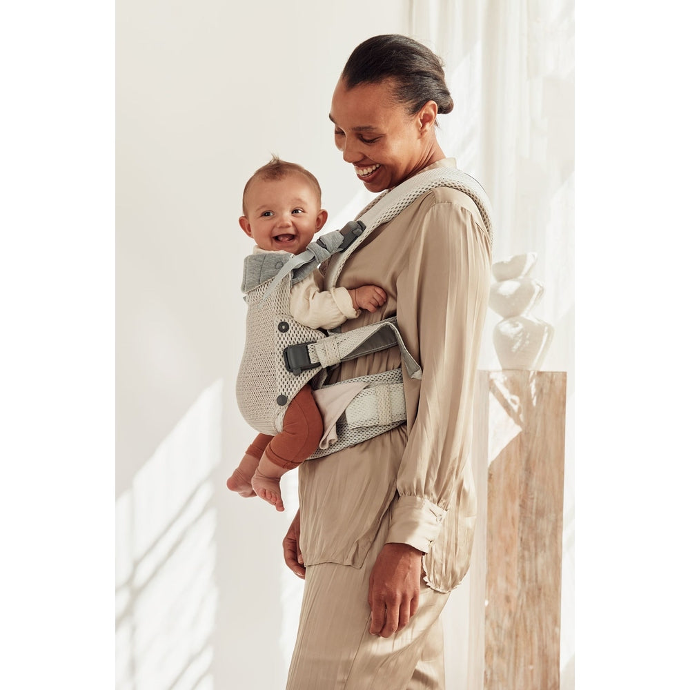 BabyBjörn Harmony baby carrier in beige, parent wearing baby on chest with secure straps, baby smiling.