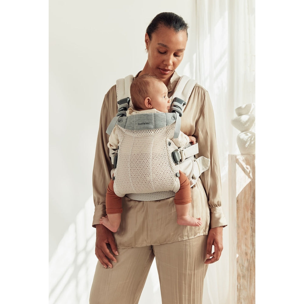 BabyBjörn Baby Carrier Harmony beige with gray straps, mom cradles baby in front carrier showing secure buckles.