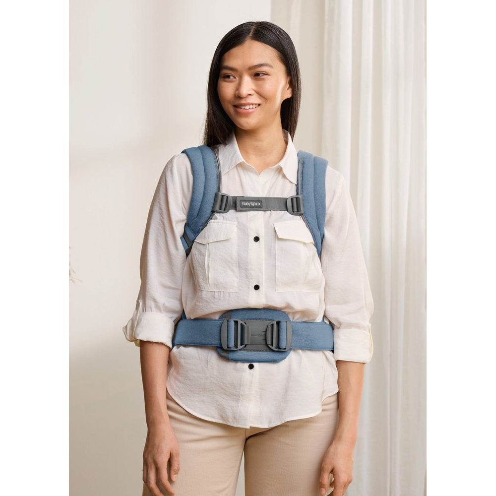 BabyBjörn Baby Carrier Harmony in blue on a smiling woman, showing padded shoulders and wide waist belt.