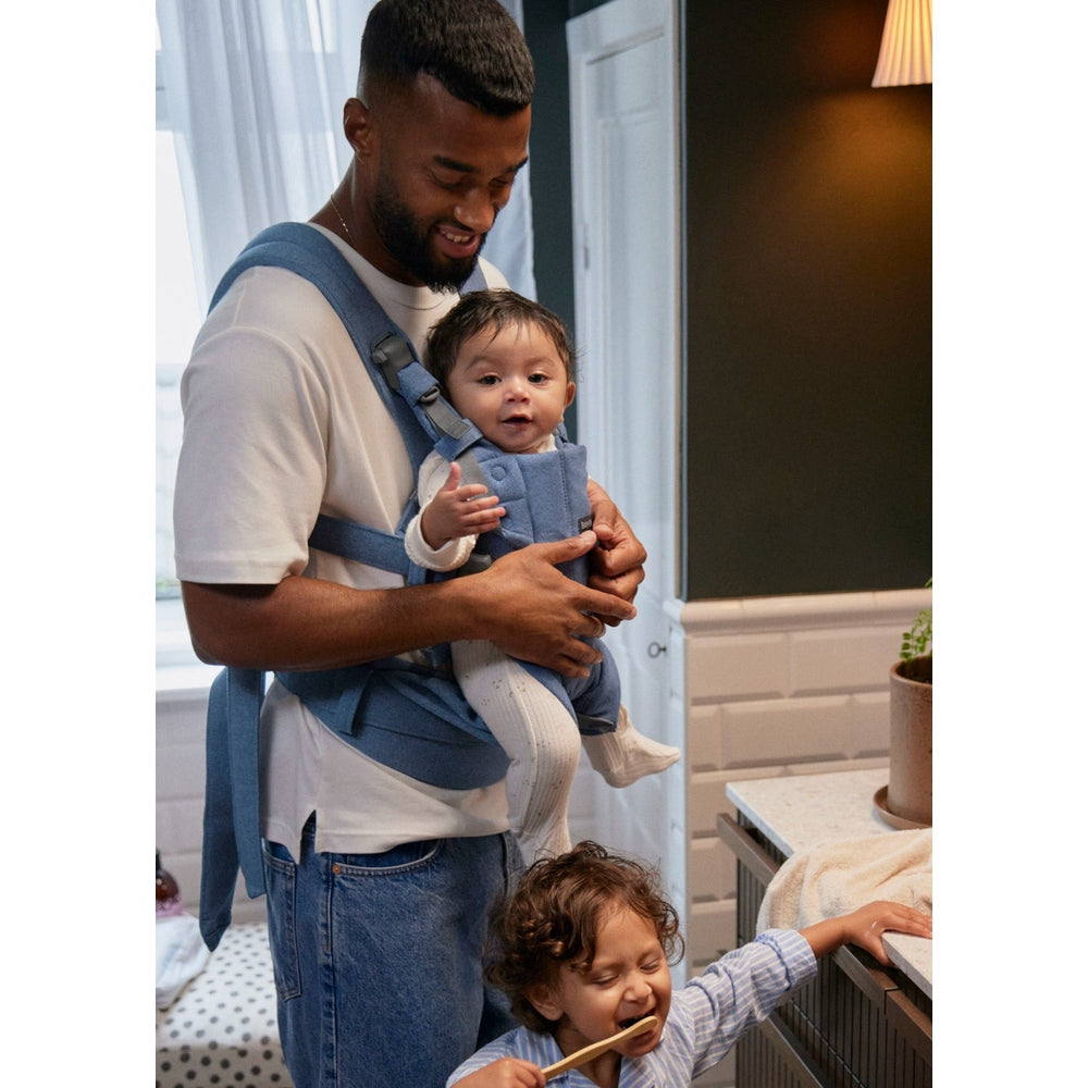 Dad wearing a blue Baby Björn Baby Carrier Harmony cradles a happy baby, showcasing secure ergonomic straps in use.