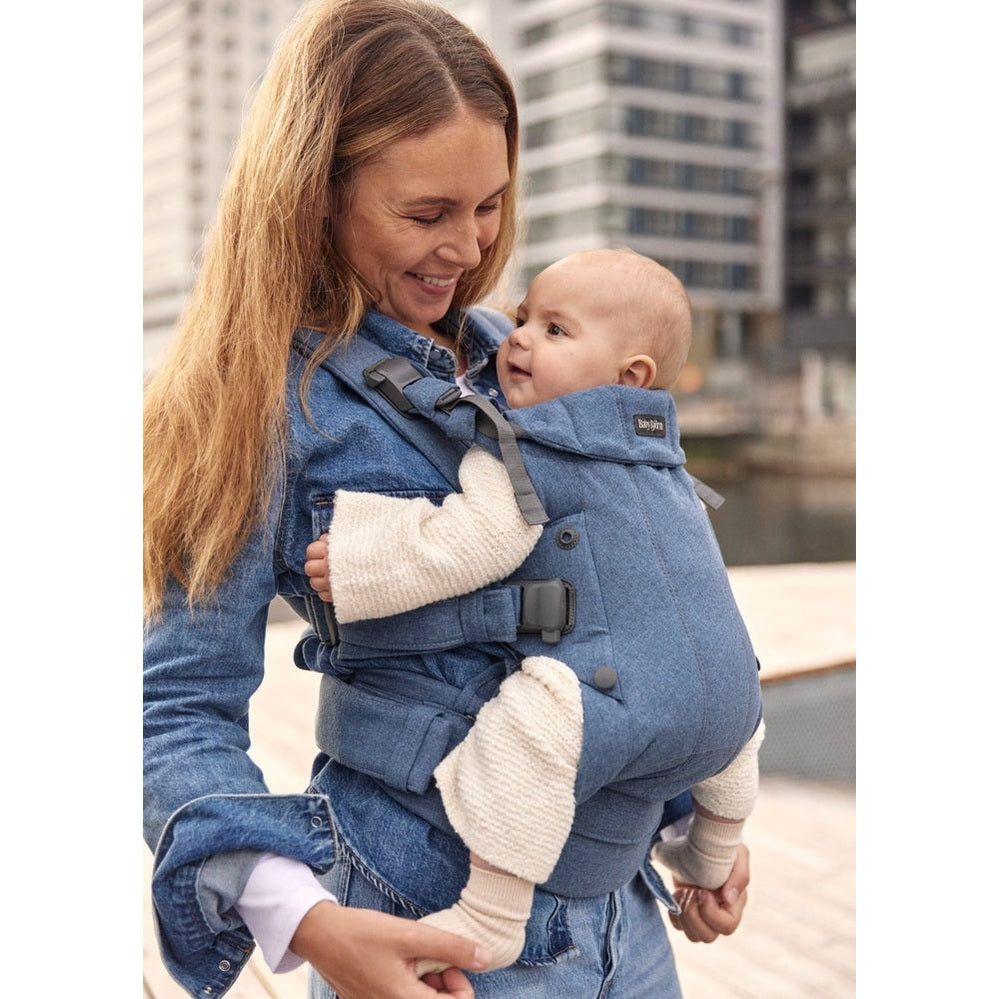 BabyBjörn Baby Carrier Harmony in blue denim worn by smiling mom carrying baby against a city backdrop.