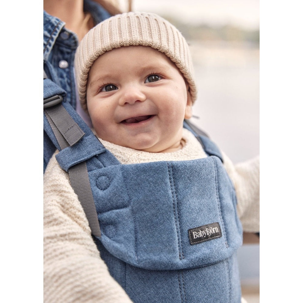 BabyBjörn Harmony baby carrier in blue denim, baby smiling while being securely carried close by a caregiver.