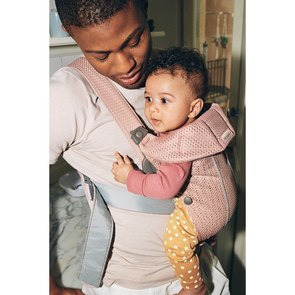 BabyBjörn Baby Carrier Mini in pink mesh, parent buckles chest strap as baby sits facing inward.
