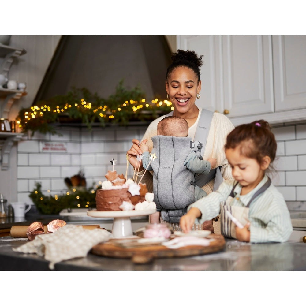 BabyBjörn Baby Carrier Mini in gray worn by smiling mom as she bakes cake with baby secured.