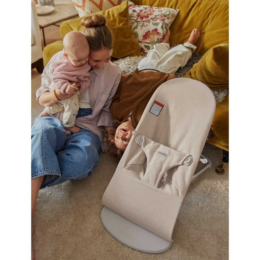 BabyBjörn Bouncer Bliss beige in a cozy living room, mother cuddling a baby while a toddler plays nearby.