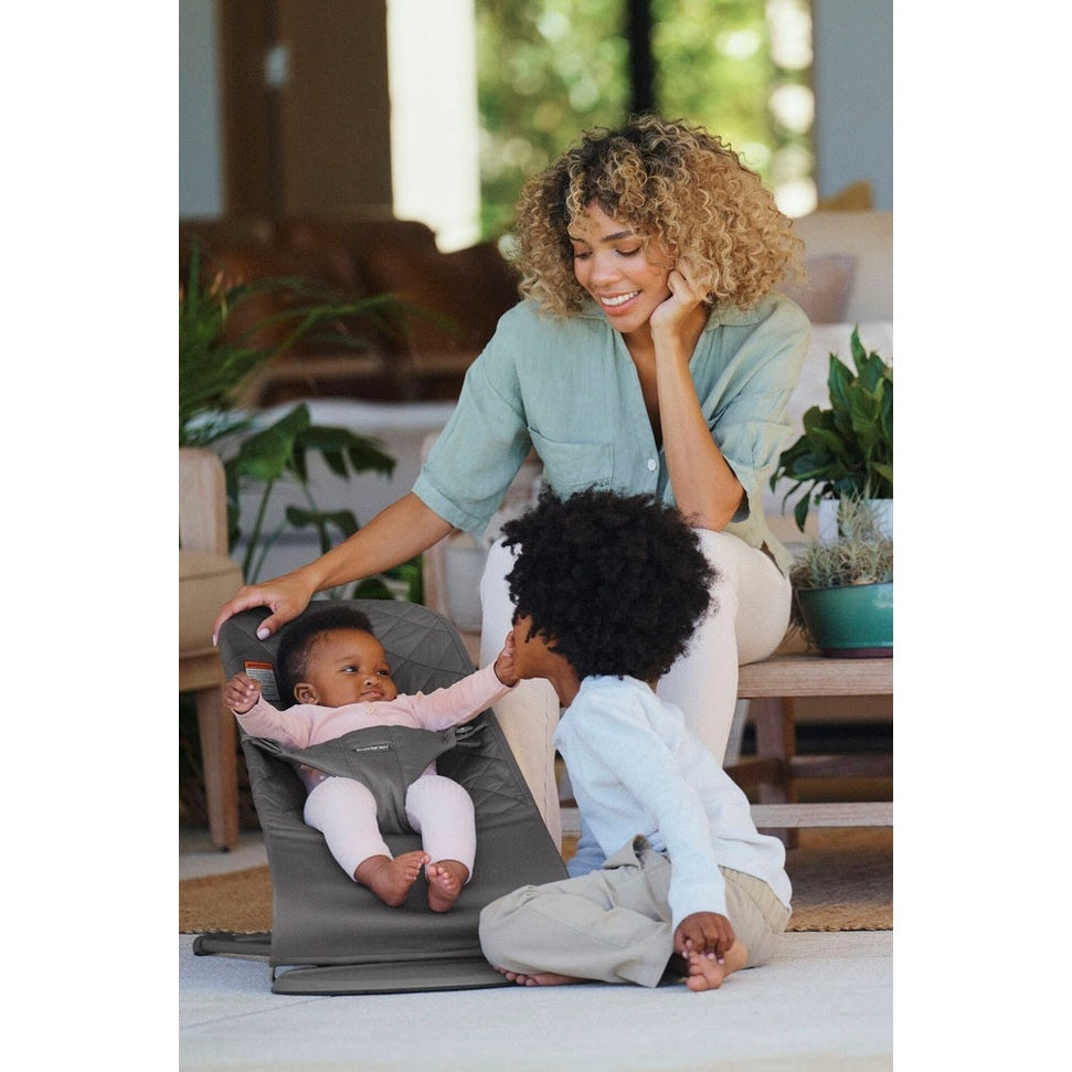 BabyBjörn Bouncer Bliss gray infant bouncer; baby secured in the seat while mom smiles nearby and toddler interacts.