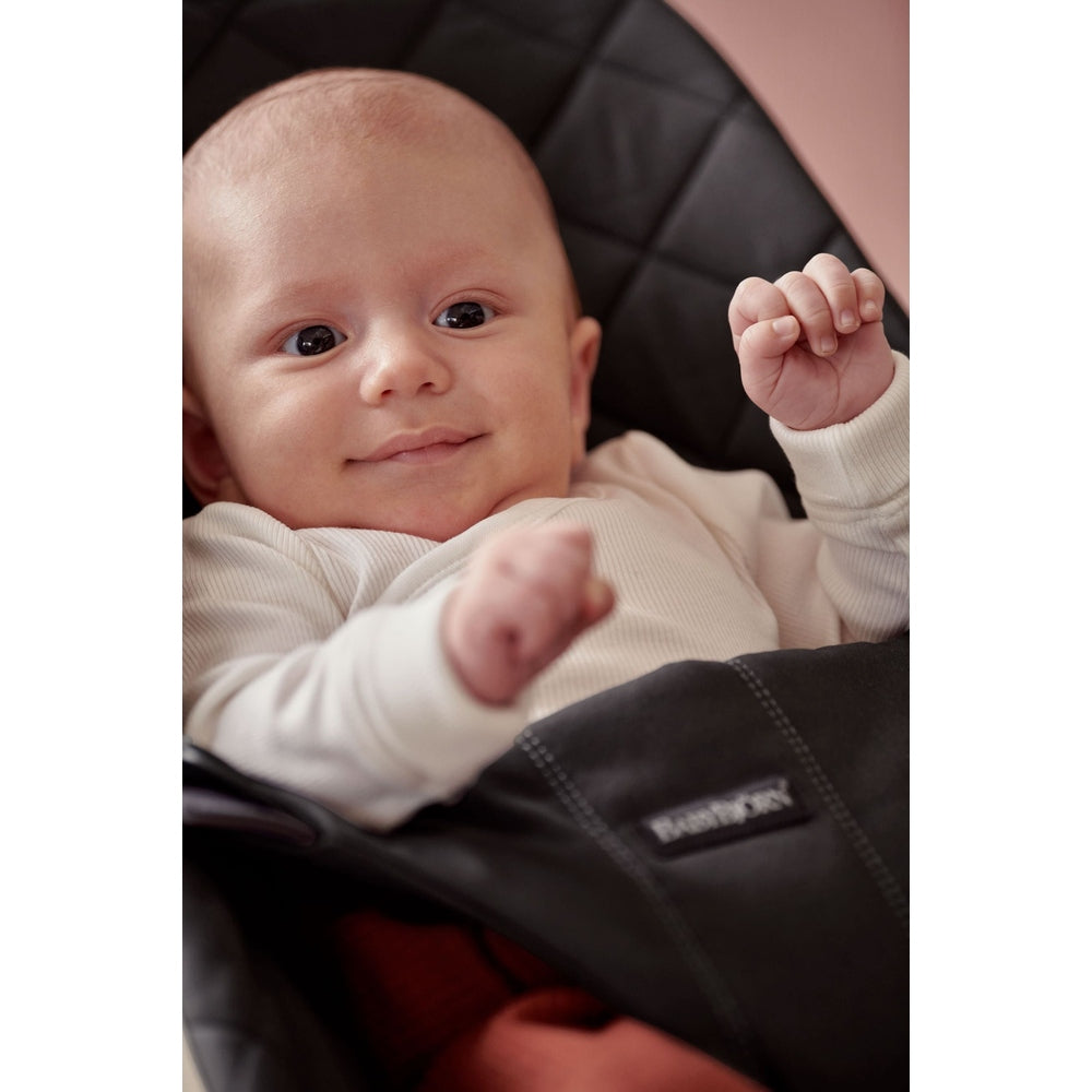 BabyBjörn Bouncer Bliss in black; smiling baby in a white onesie sits securely in the padded seat.