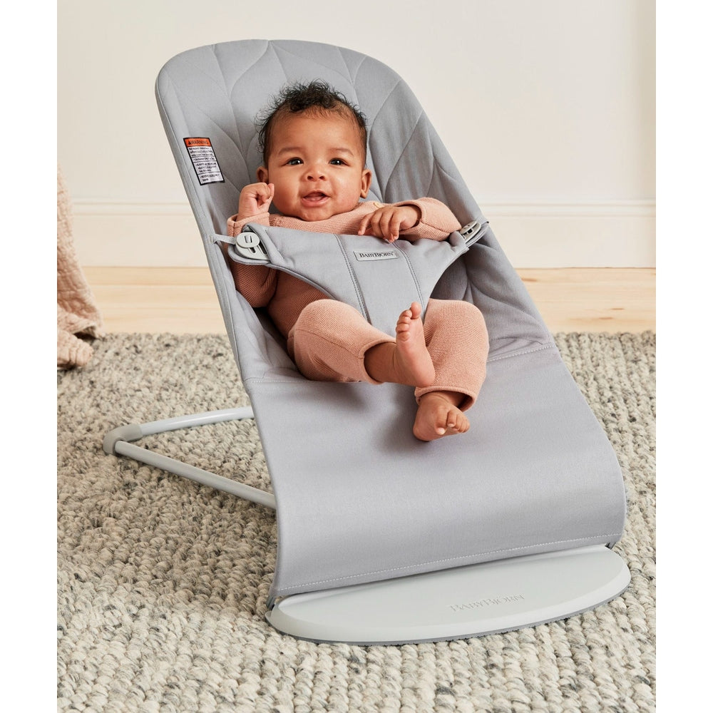 BabyBjörn Bouncer Bliss in soft grey; smiling baby sits securely in the reclined fabric seat using the 5-point harness.