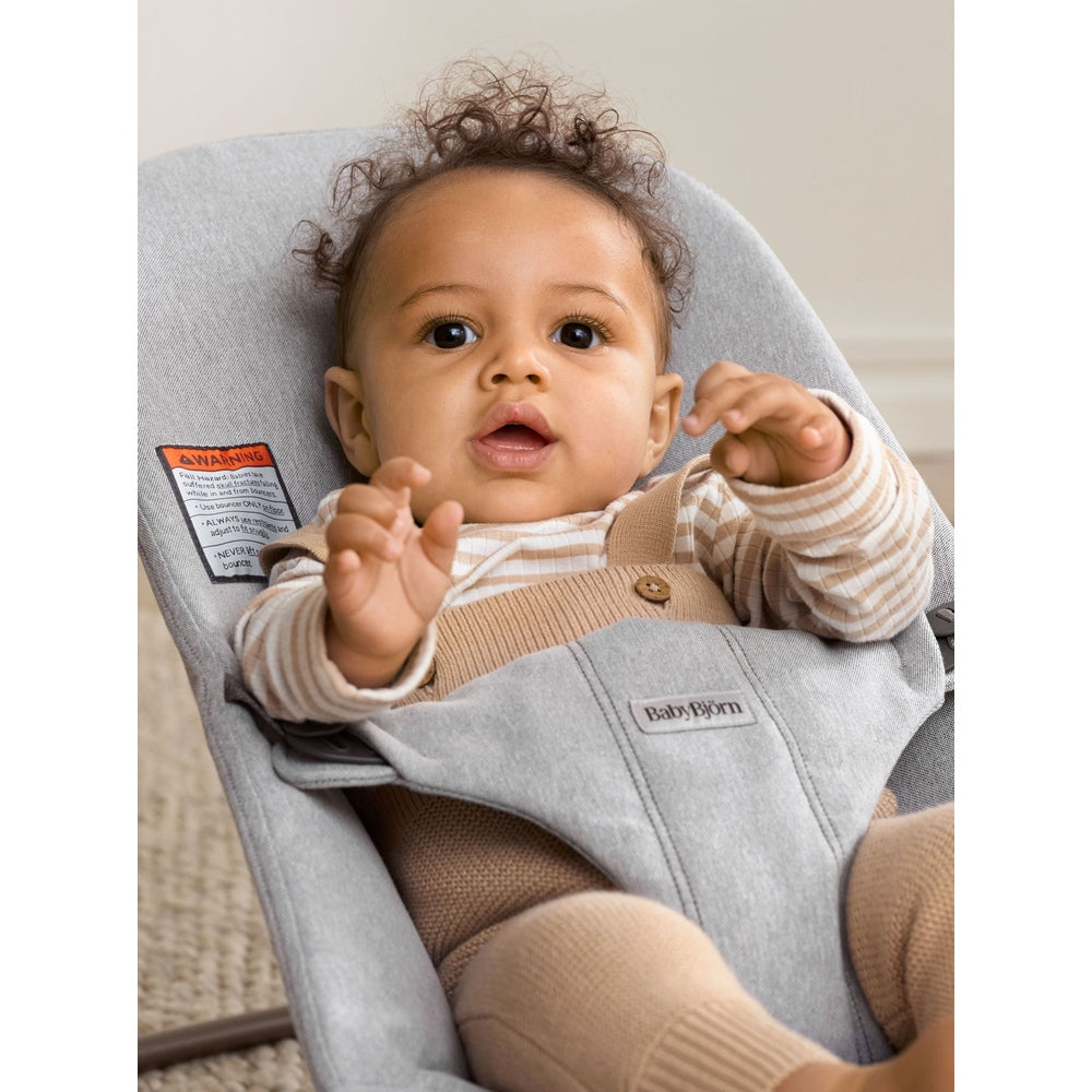 BabyBjörn Bouncer Bliss in grey fabric, baby secured in beige harness while reaching hands.