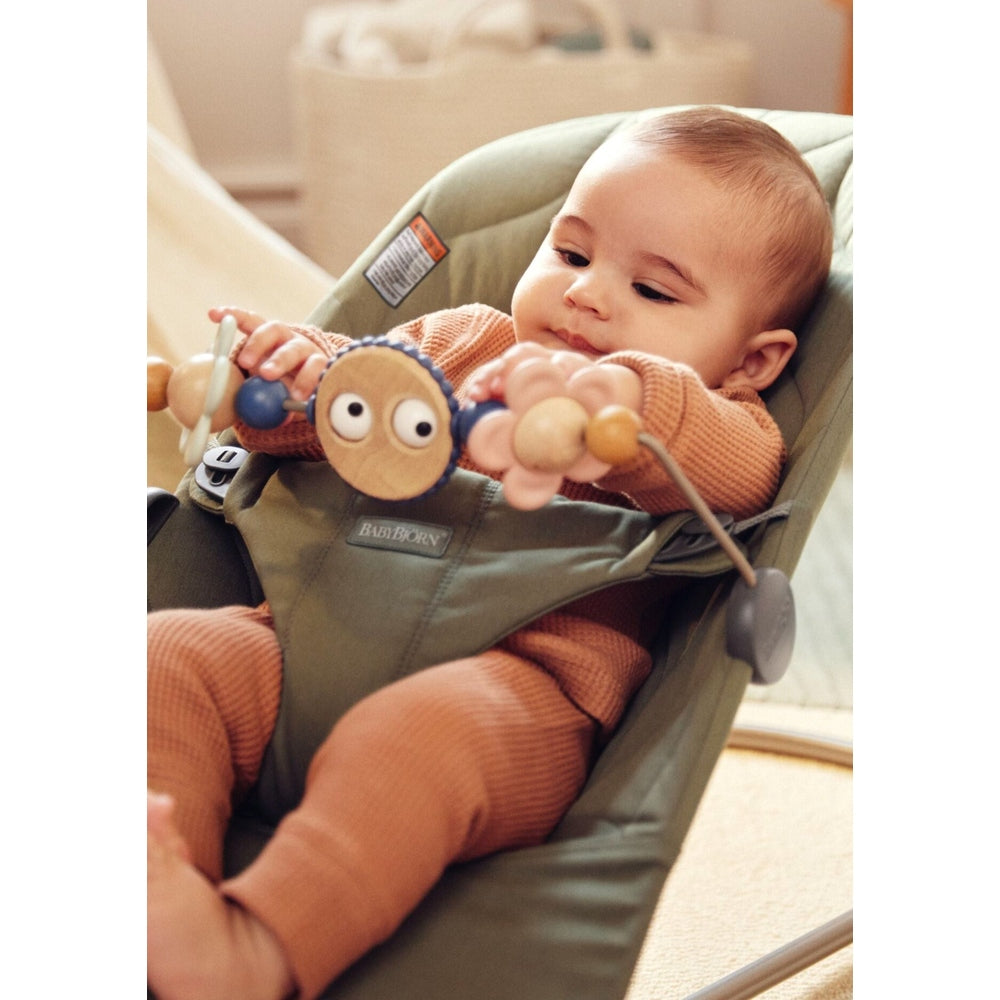 BabyBjörn Bouncer Bliss in green fabric; baby plays with a wooden bead arch while seated and relaxed.