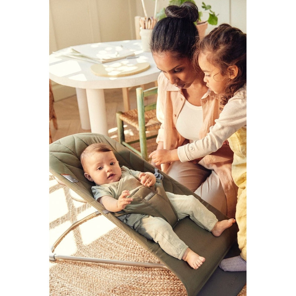 BabyBjörn Bouncer Bliss in green; baby reclines secured in harness while caregiver smiles nearby.