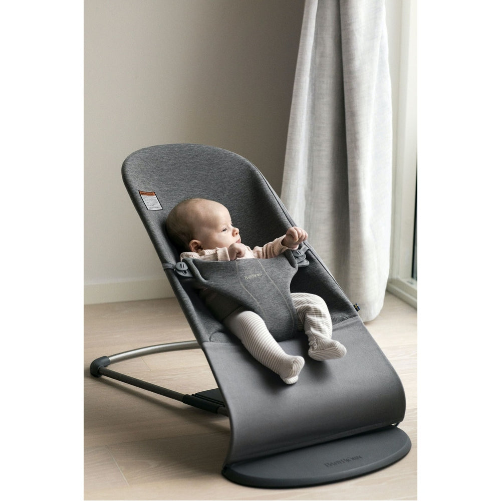 BabyBjörn Bouncer Bliss in gray, a baby relaxing and reaching forward in a bright home setting.