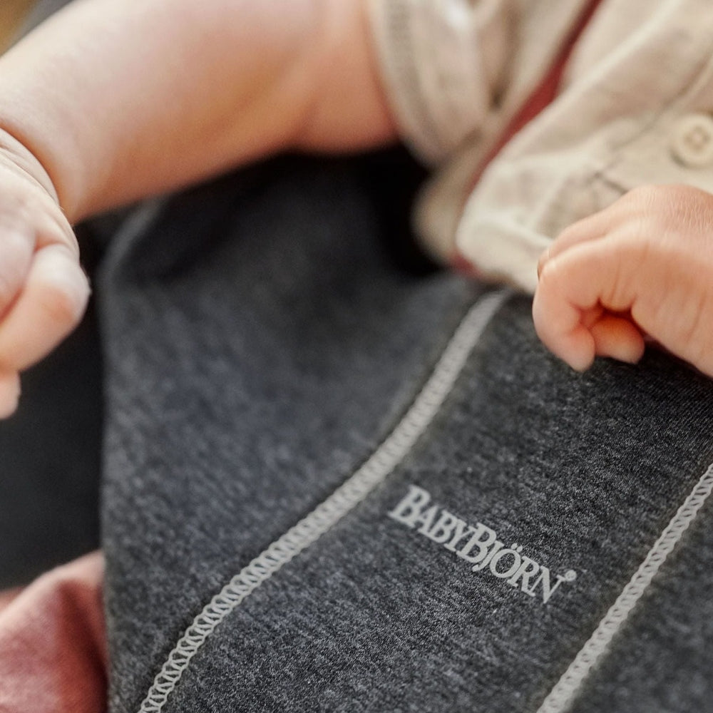 BabyBjörn Bouncer Bliss in dark gray fabric; a baby grips the soft edge, showing plush comfort and sturdy stitching.