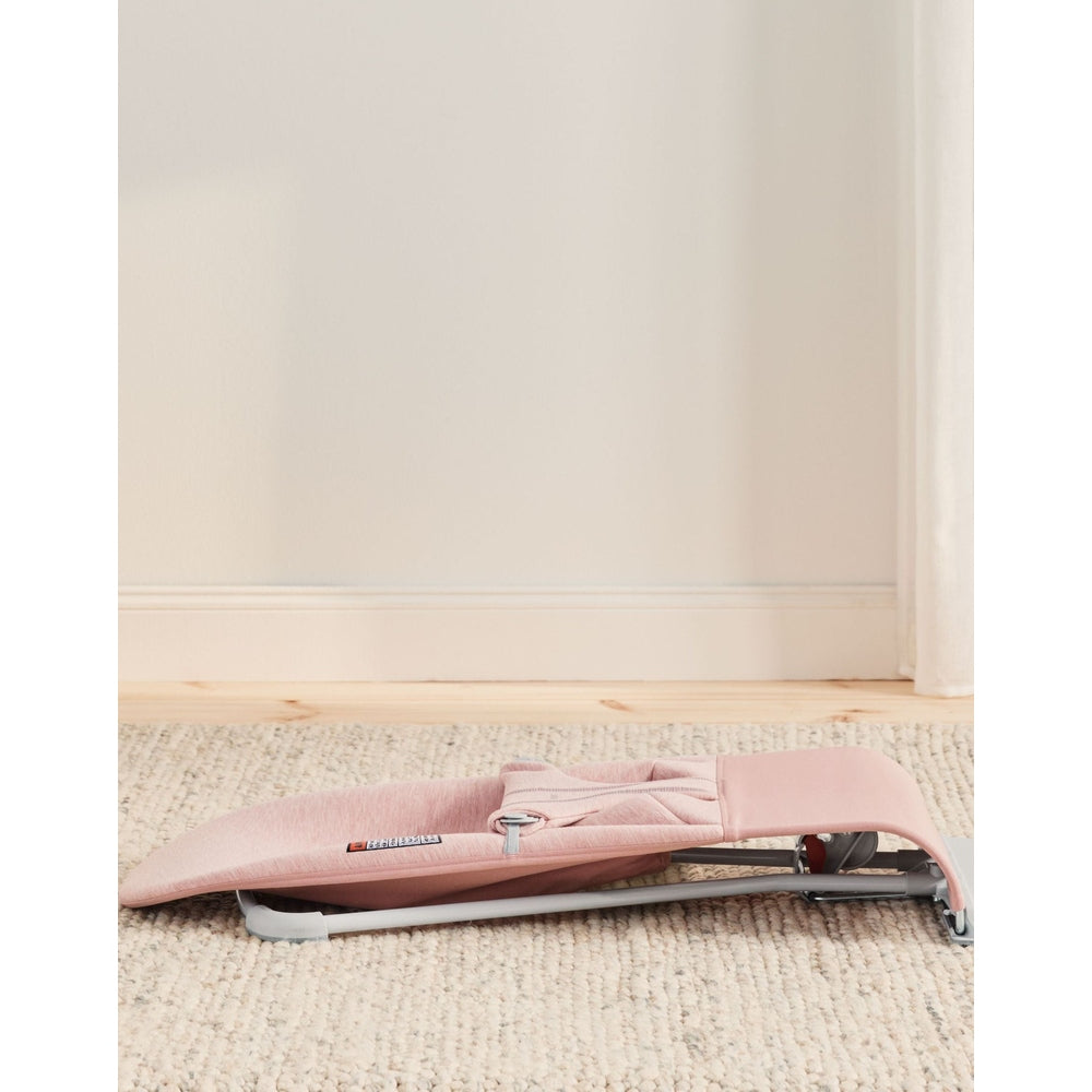 BabyBjörn Bouncer Bliss in soft pink, folded flat on a beige rug with its lightweight frame exposed.