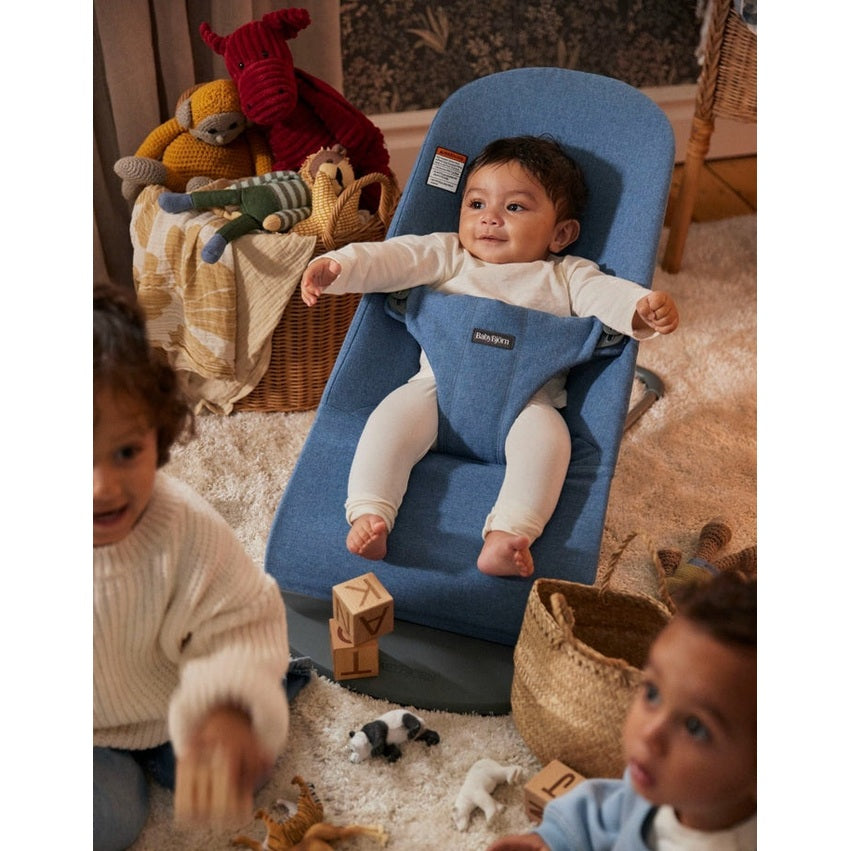 BabyBjörn Bouncer Bliss blue fabric baby rocker with a baby sitting upright and arms out, toys nearby.