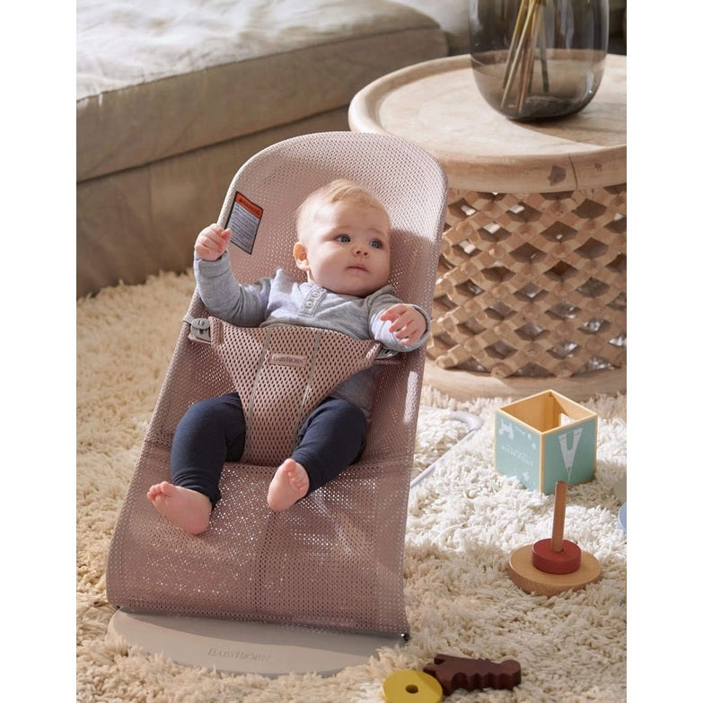 BabyBjörn Bouncer Bliss in pink mesh, baby strapped in and reaching hands up in a cozy living room.