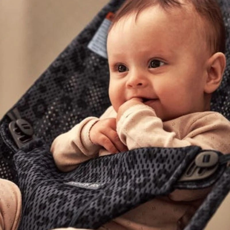 BabyBjörn Bouncer Bliss in dark grey mesh, baby calmly smiles and sucks fingers while seated.