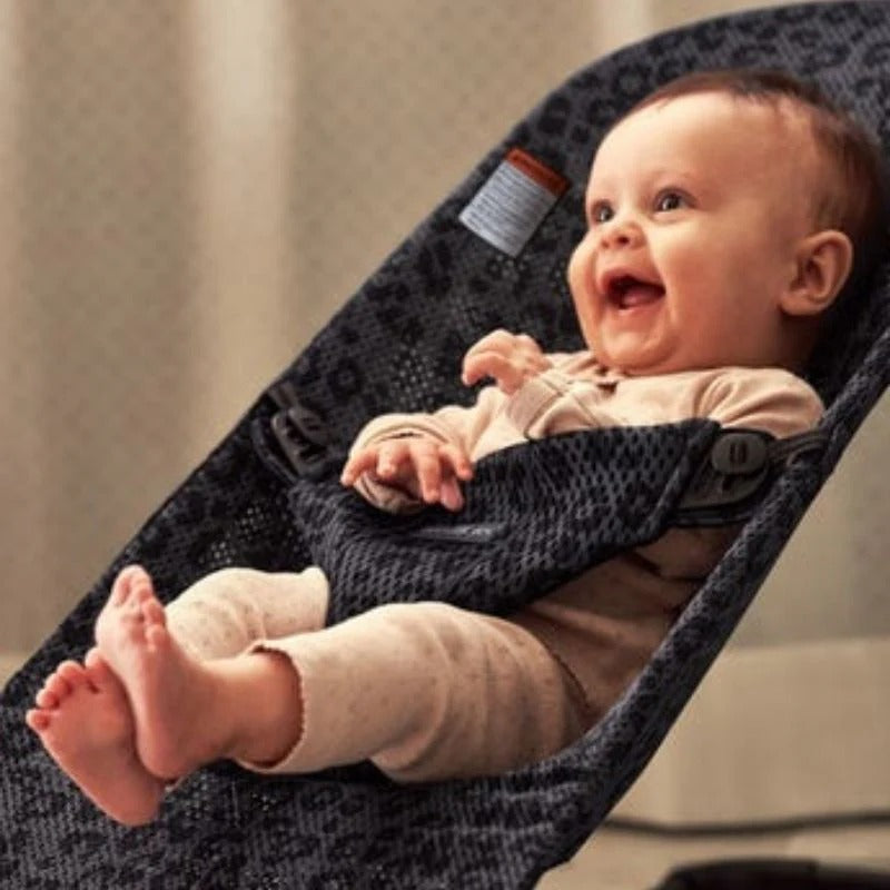 BabyBjörn Bouncer Bliss in dark patterned fabric with BABYBJÖRN tag; smiling baby reclines securely in the rocker.