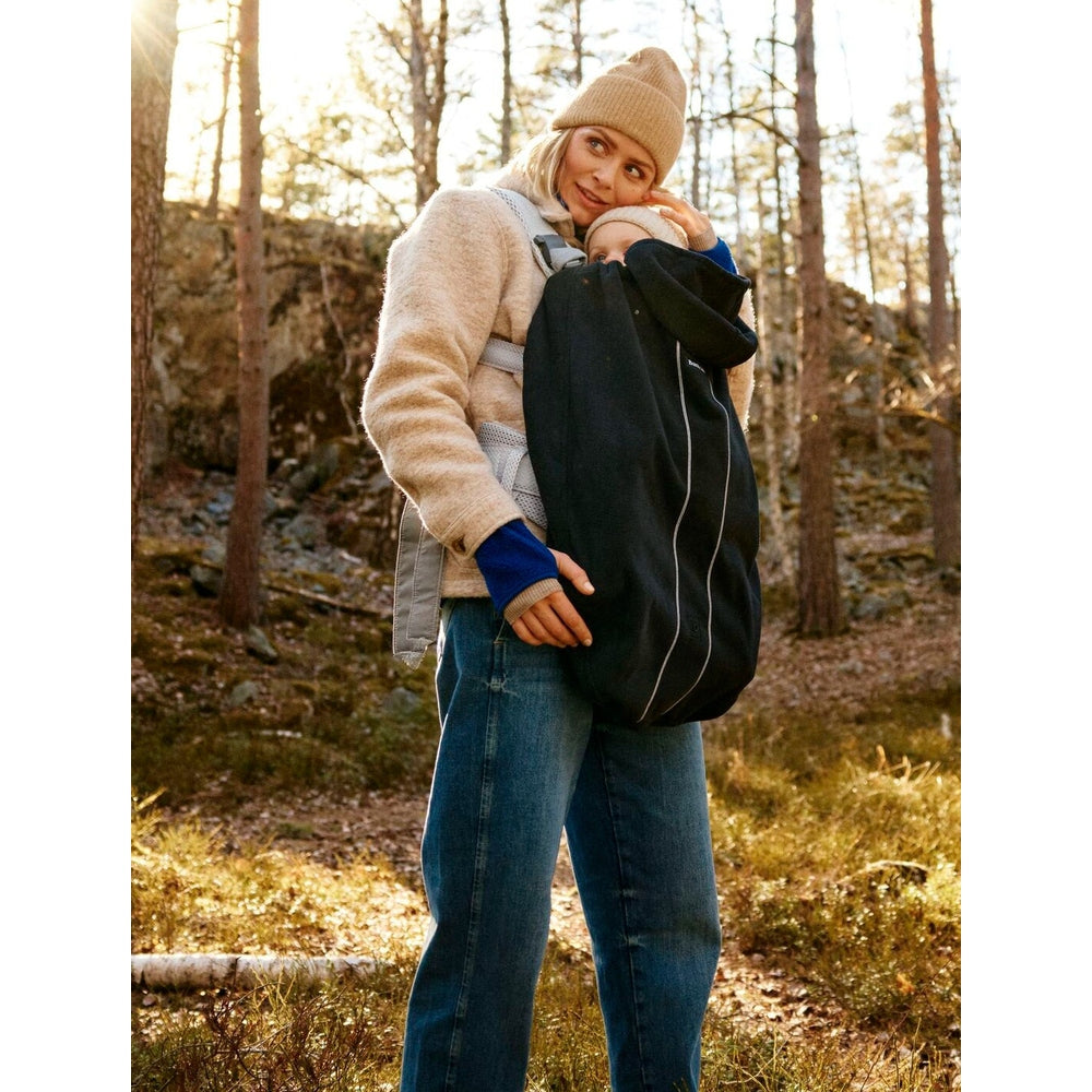 Mother outdoors in a forest, wearing a beige beanie, carries baby in a black BABY BJÖRN Cover for Baby Carrier.