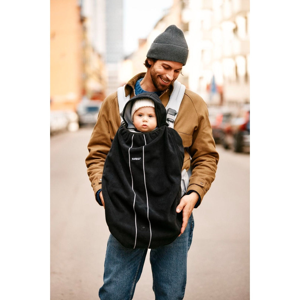 BabyBjörn Cover for Baby Carrier worn by a smiling dad carrying a baby on a city street in a black cover with white piping