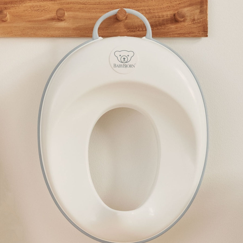 BabyBjörn Toilet Trainer in white with gray trim, hanging on a wooden wall hook.