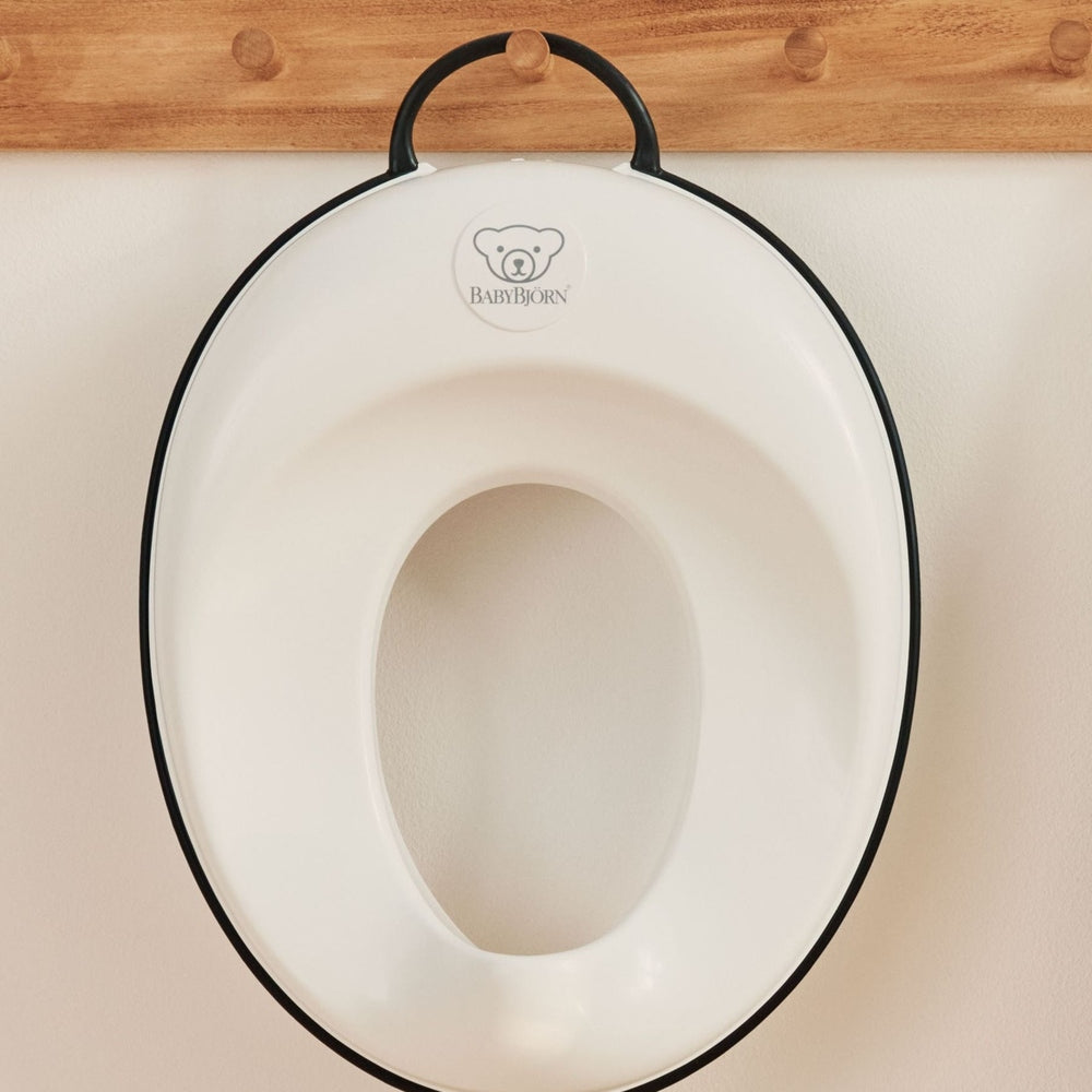 BabyBjörn Toilet Trainer, white with black edge, hanging on a wooden rack in a nursery.