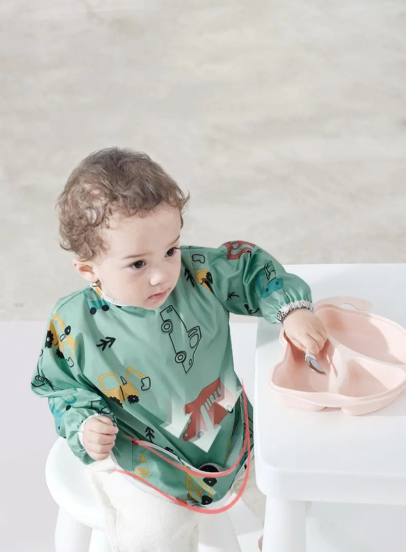 BC Babycare Long Sleeve Bib in green car print with front pocket, toddler wears it at table to catch spills.