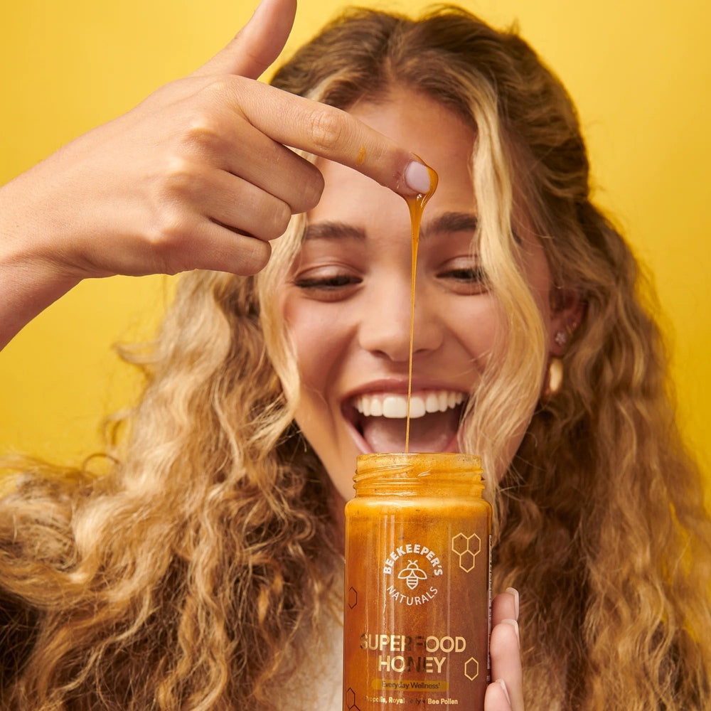 Beekeeper's Naturals Superfood Honey 330g; joyful woman drips golden honey from her finger into the jar.
