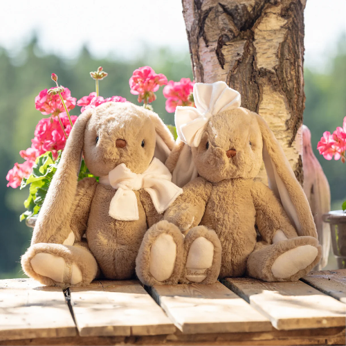 Bukowski Bears Bunny Patrick stuffed animals pair, beige with cream bows, sitting together on a wooden deck near pink flowers.