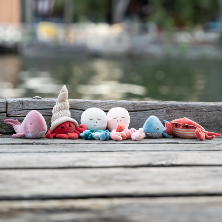 Bukowski Bears Hermit The Crab stuffed animal among colorful sea plushies on a weathered dock beside calm water.