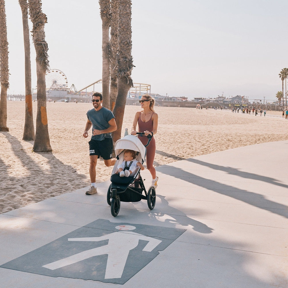 CYBEX Avi Spin All Terrain Stroller on a beach boardwalk, mom pushing with baby as dad jogs nearby.