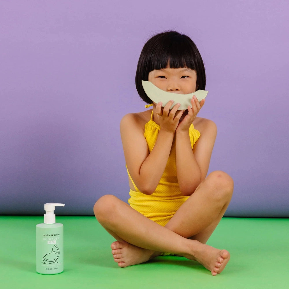 Dabble & Dollop All-Natural Shampoo, Body Wash & Bubbles Honeydew bottle beside a smiling girl in a yellow swimsuit.