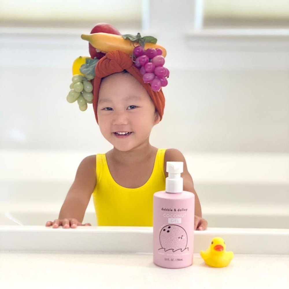 Dabble & Dollop All-Natural Coconut shampoo bottle beside a smiling child in a yellow swimsuit, ready for bath.