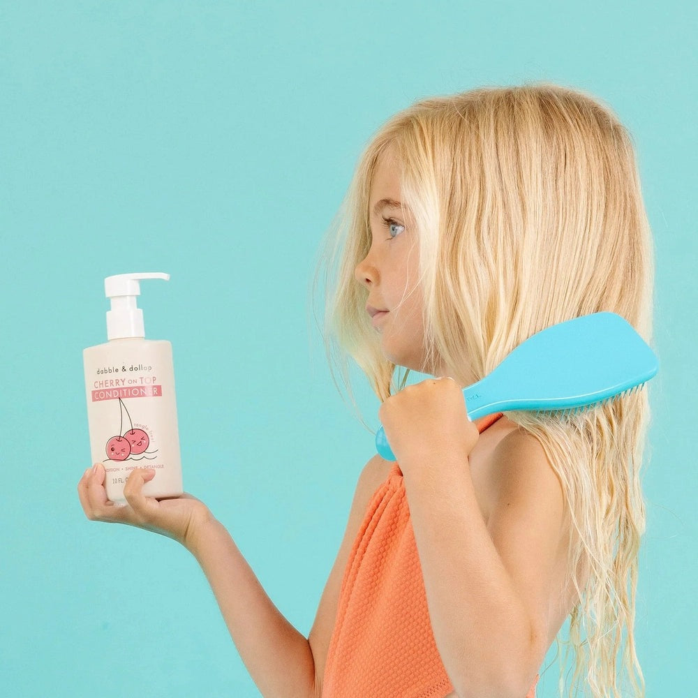 Dabble & Dollop Hair Conditioner Cherry on Top bottle with pump held by girl, blue detangler comb nearby.