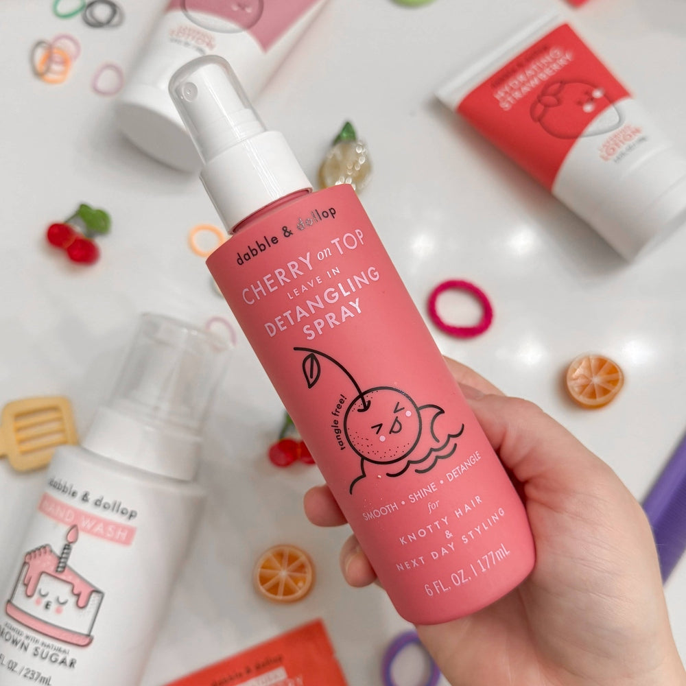 Dabble & Dollop Cherry on Top leave-in detangling spray in a pink bottle held in hand amid colorful beauty products