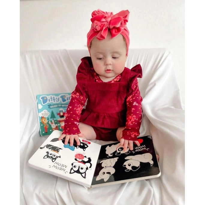 Baby girl in a red outfit sits on a white couch with Ditty Bird Sound Book Black and White Animals USB-C Rechargeable, flipping pages.
