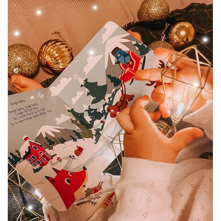 Ditty Bird Sound Book Christmas Songs USB-C Rechargeable being opened by a child beside twinkling holiday lights.