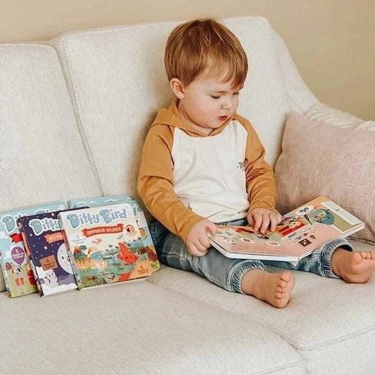 Ditty Bird Sound Book Dinosaur Sounds USB-C Rechargeable; toddler sits on a sofa tapping the colorful pages.