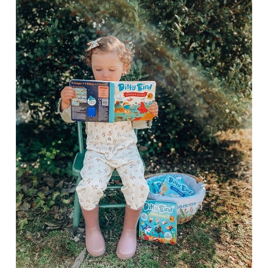 Ditty Bird Sound Book Dinosaur Sounds USB-C rechargeable, a child reads it outdoors on a green chair.