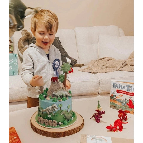 Ditty Bird Dinosaur Sounds educational toy box on a table beside a delighted child with colorful dinosaur figurines.