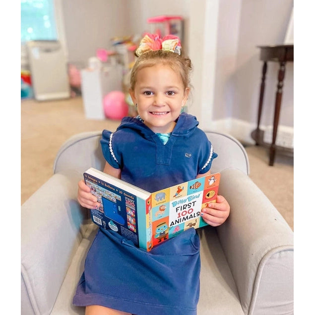 Little girl in a blue dress sits in a chair reading the Ditty Bird Sound Book First 100 Animals USB-C Rechargeable, smiling at the colorful pages.