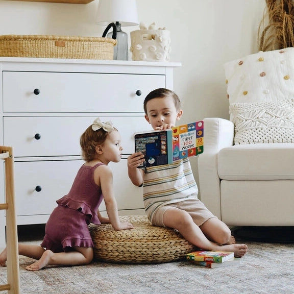 Ditty Bird Sound Book First 100 Words USB-C Rechargeable being read by two kids on a woven pouf in a cozy room.