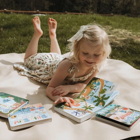A girl on a blanket outdoors taps a colorful Ditty Bird Sound Book Fun Alphabet USB-C Rechargeable, learning letters.