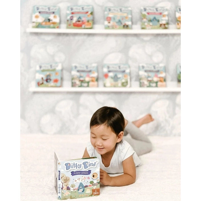 Ditty Bird Sound Book Funny Songs USB-C Rechargeable; a toddler on a bed reaches for the colorful book, with Ditty Bird toys on shelves behind.