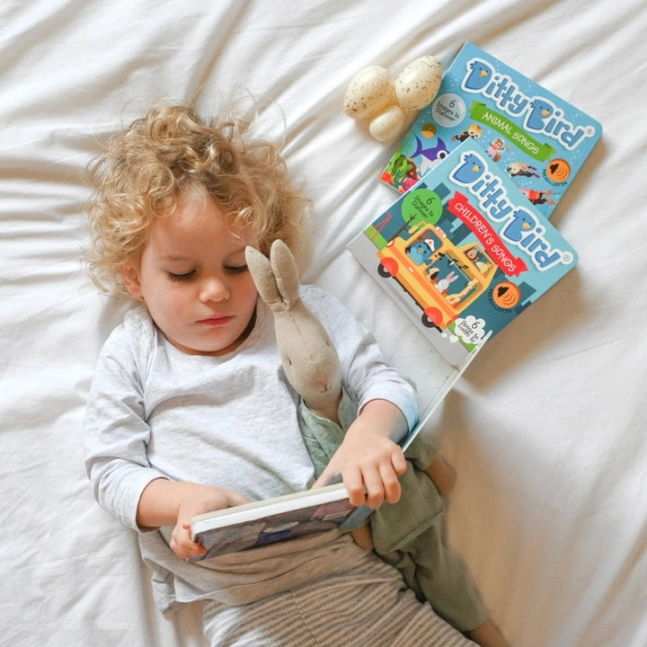 Ditty Bird Sound Book Funny Songs USB-C Rechargeable, toddler reads on bed with plush bunny and two Ditty Bird books.