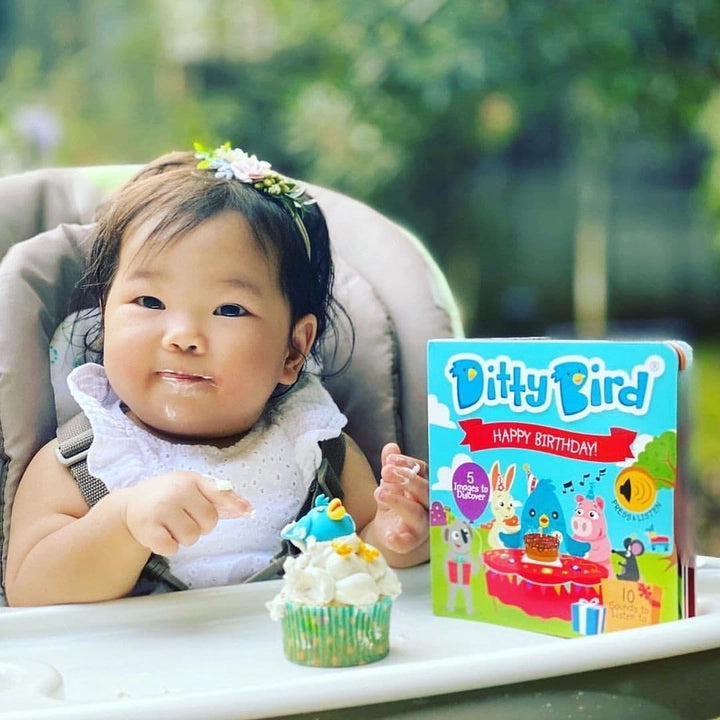 Ditty Bird Sound Book Happy Birthday USB-C Rechargeable beside a smiling baby in a high chair with a cupcake on the tray.