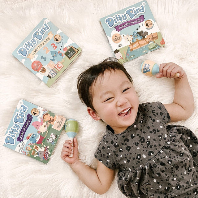 Ditty Bird Sound Book Learning Songs USB-C Rechargeable; happy baby on a white rug grips a blue Ditty Bird toy beside three learning books.