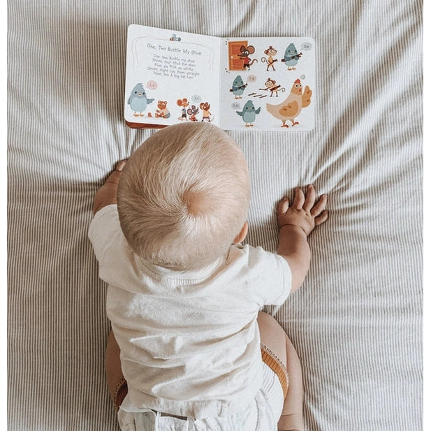 Ditty Bird Sound Book Learning Songs USB-C Rechargeable; baby sits on a striped bed reading the colorful pages.