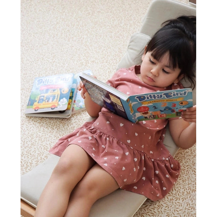 Ditty Bird Sound Book Learning Songs USB-C Rechargeable toy; a girl in a pink dress reads it on a lounge chair.