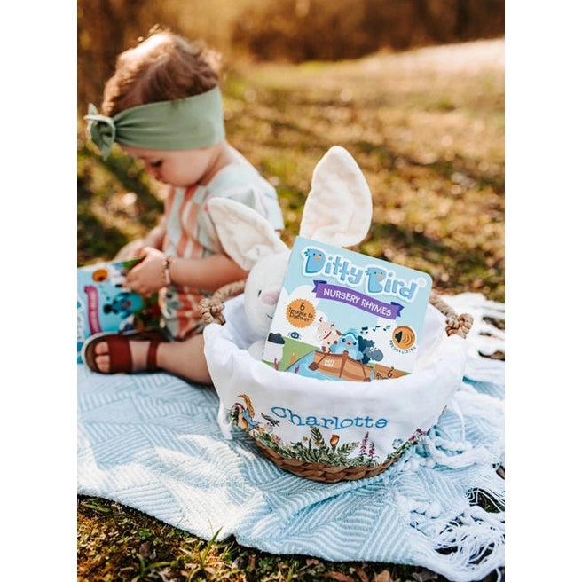 Ditty Bird Sound Book Nursery Rhymes USB-C Rechargeable toddler sits on a blanket outdoors beside a basket of Ditty Bird.